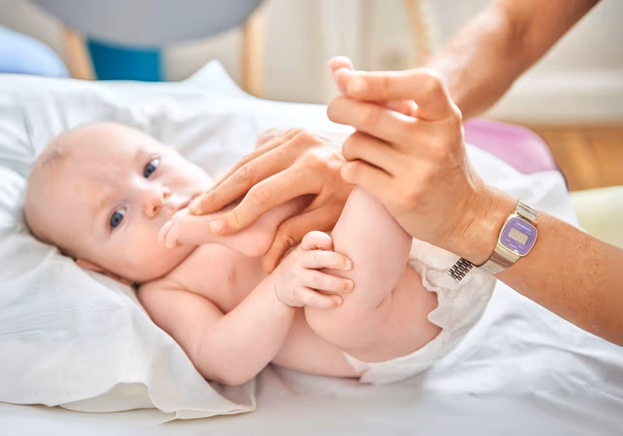 foto di un neonato su un letto con lenzuola bianche. le mani di una donna fanno una presa osteopatica su neonato. La donna porta un orologio sul polso della mano sinistra